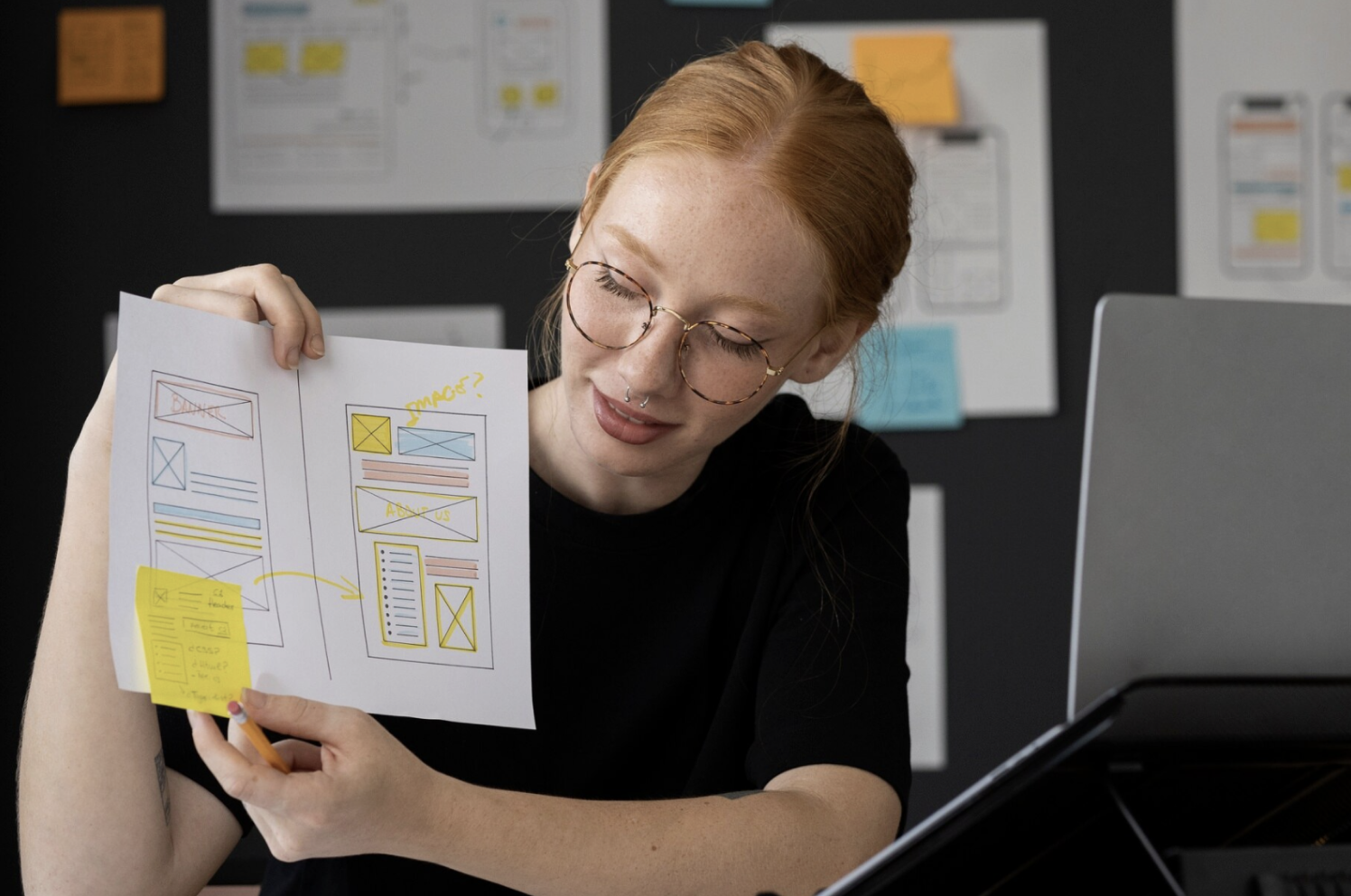 Diseñadora mostrando dos wireframes dibujados a mano frente a un muro de bocetos de interfaz.
